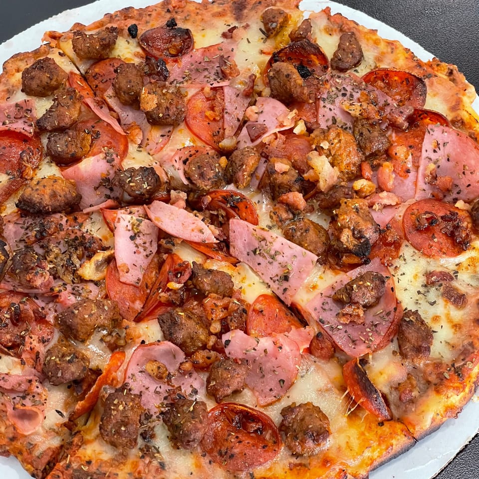 Close-up of a loaded pizza topped with meatballs, pepperoni, ham, tomatoes, melted cheese, and herbs on a white plate.