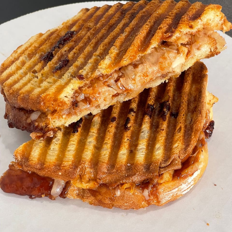 Close-up of a golden grilled sandwich with layered flaky bread, melted cheese, sliced meat, and glossy sauce on a white plate.